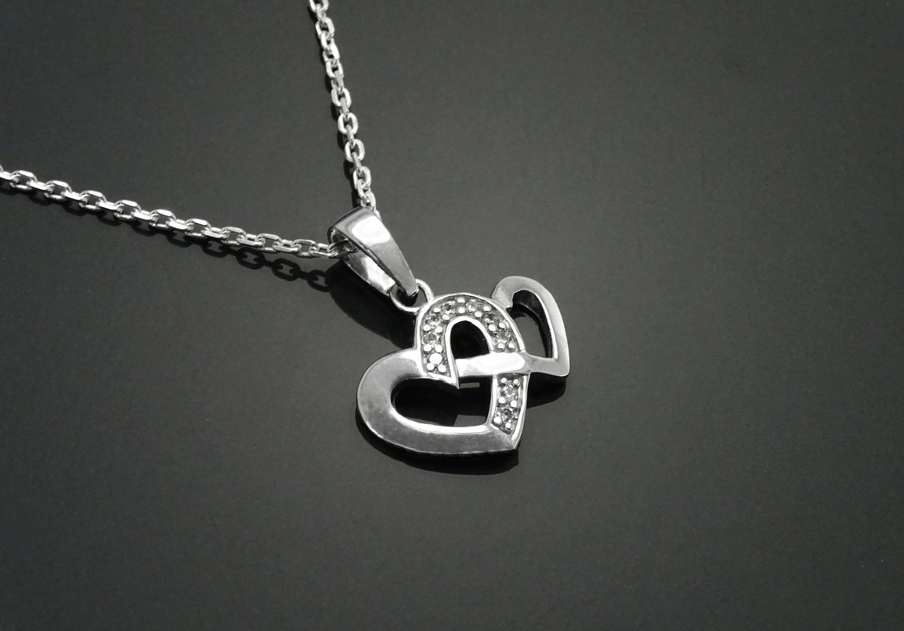 Un collier en argent avec un pendentif en forme de deux cœurs entrelacés. Les cœurs sont ornés de petites pierres scintillantes.