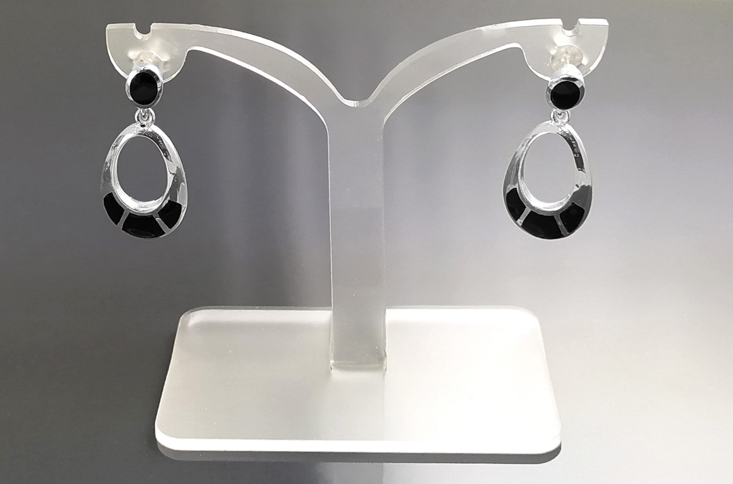 Une paire de boucles d'oreilles ovales pendantes en argent 925 avec des accents noirs de forme géométrique. Les boucles d'oreilles sont présentées sur un support blanc.