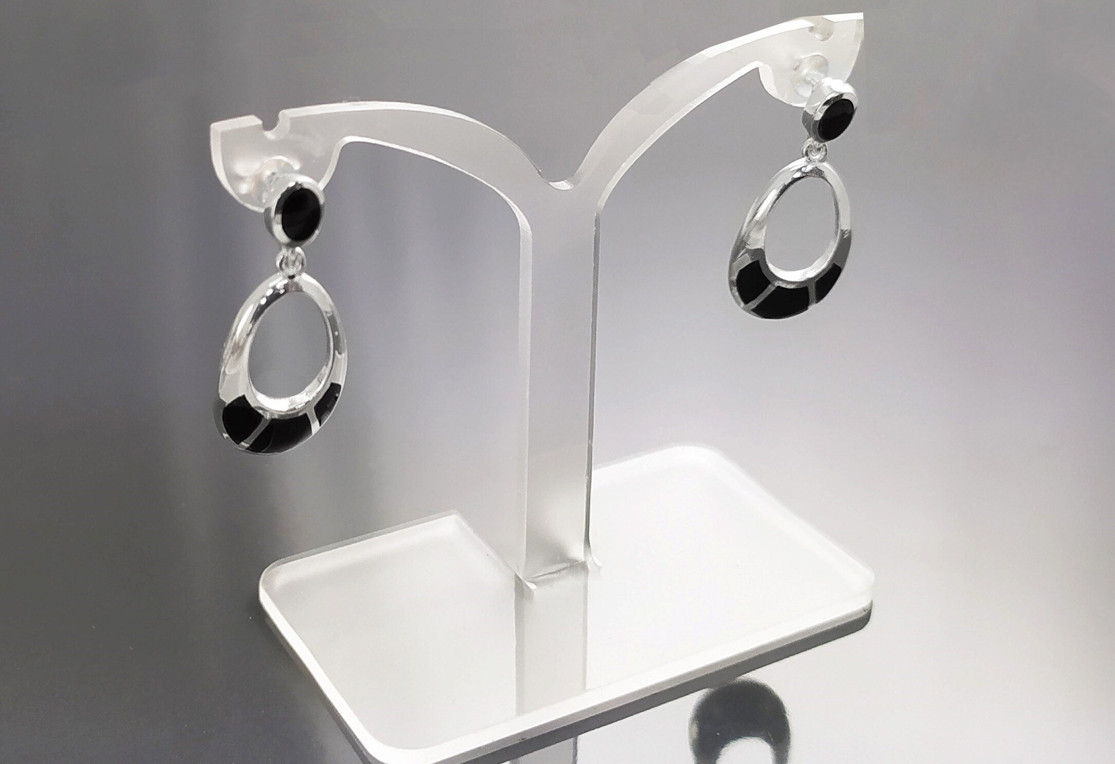 Une paire de boucles d'oreilles ovales pendantes en argent 925 serties avec des pierres noires en onyx. Les boucles d'oreilles sont présentées sur un support en acrylique blanc.