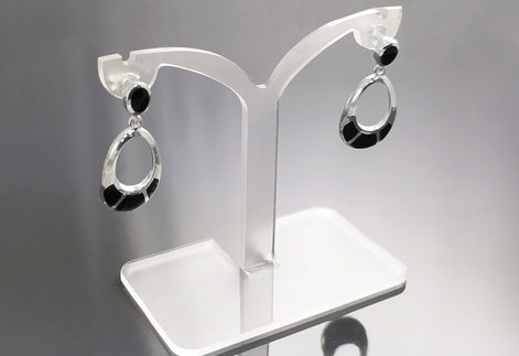 Une paire de boucles d'oreilles ovales pendantes en argent 925 serties avec des pierres noires en onyx. Les boucles d'oreilles sont présentées sur un support en acrylique blanc.