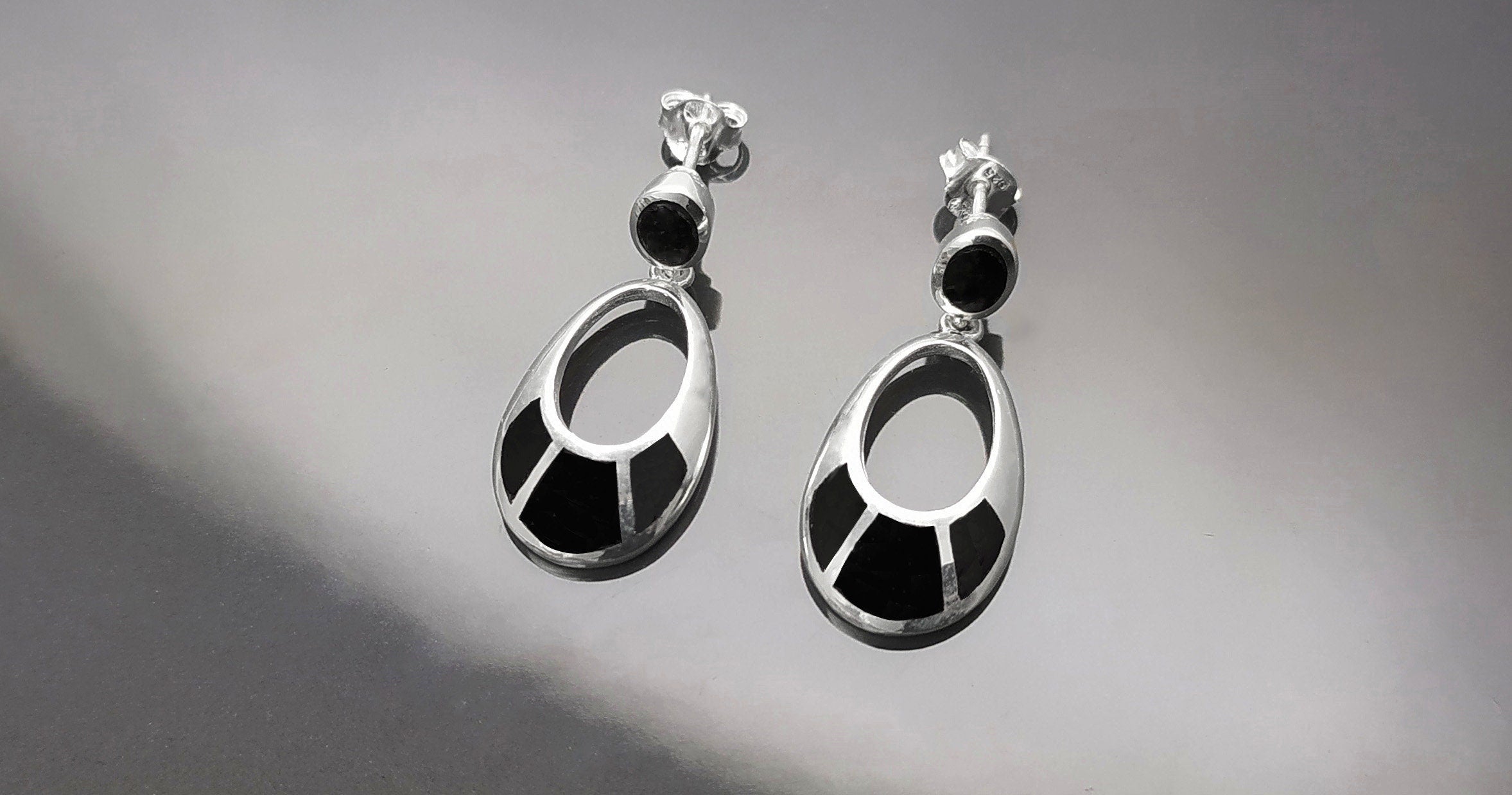 Une paire de boucles d'oreilles pendantes en argent 925 avec des onyx noirs. Les boucles d'oreilles ont un design simple, avec une petite pierre noire en haut et des plus grandes pierres noires sur la forme ovale du bas.