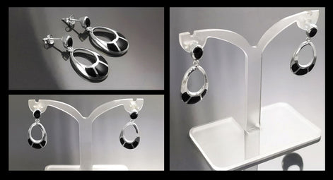 Une paire de boucles d'oreilles pendantes en argent 925 avec des pierres d'onyx noir. Les boucles d'oreilles sont de forme ovale avec un cercle découpé au centre. Les boucles d'oreilles sont sur un présentoir blanc.