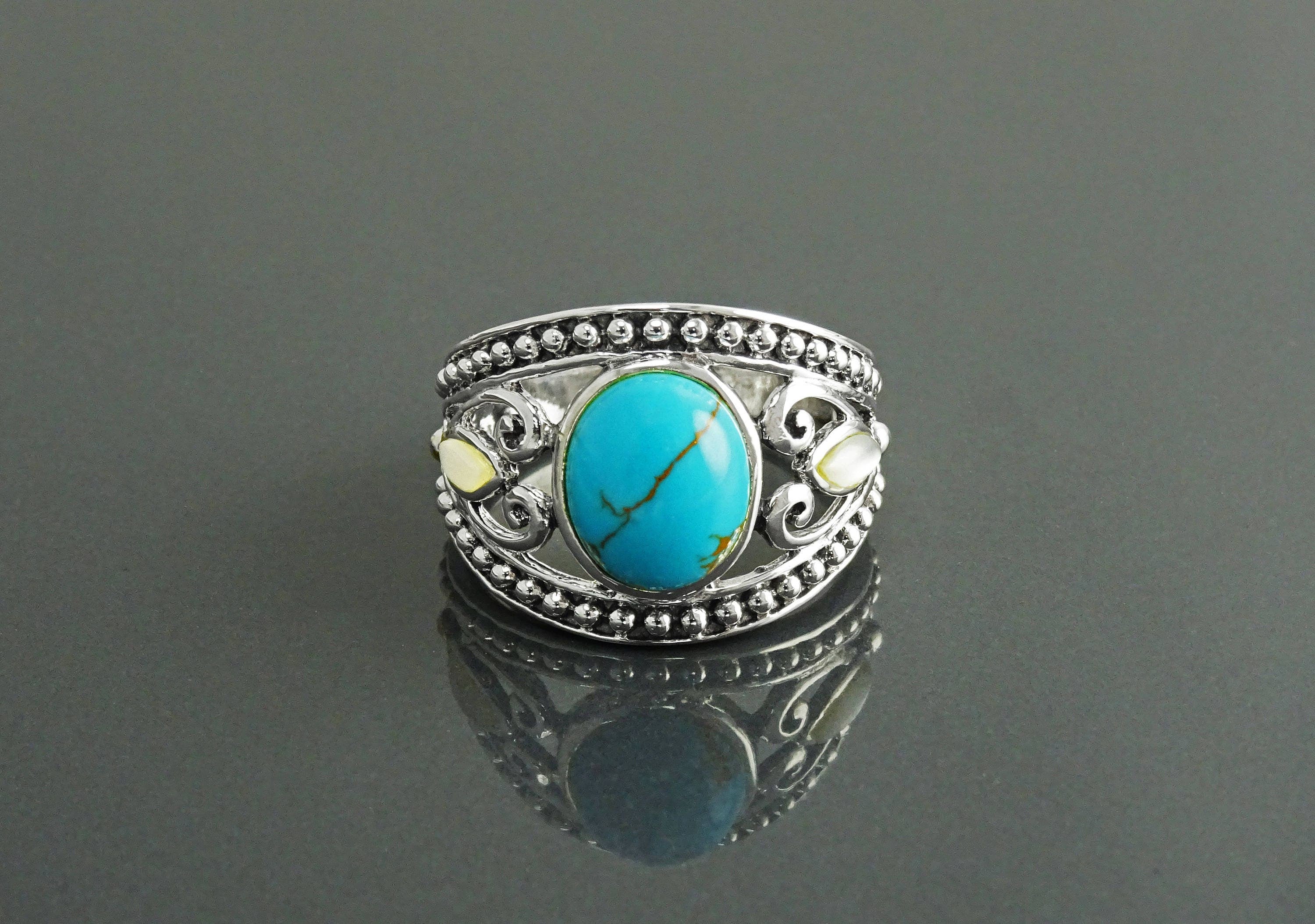 Une bague en argent 925 avec une pierre turquoise sertie dans un motif de filigrane décoratif. La bague a une bande perlée et deux petites nacres blanches de chaque côté de la pierre turquoise.