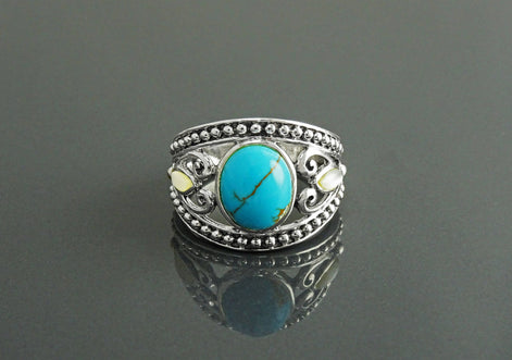 Une bague en argent 925 avec une pierre turquoise sertie dans un motif de filigrane décoratif. La bague a une bande perlée et deux petites nacres blanches de chaque côté de la pierre turquoise.