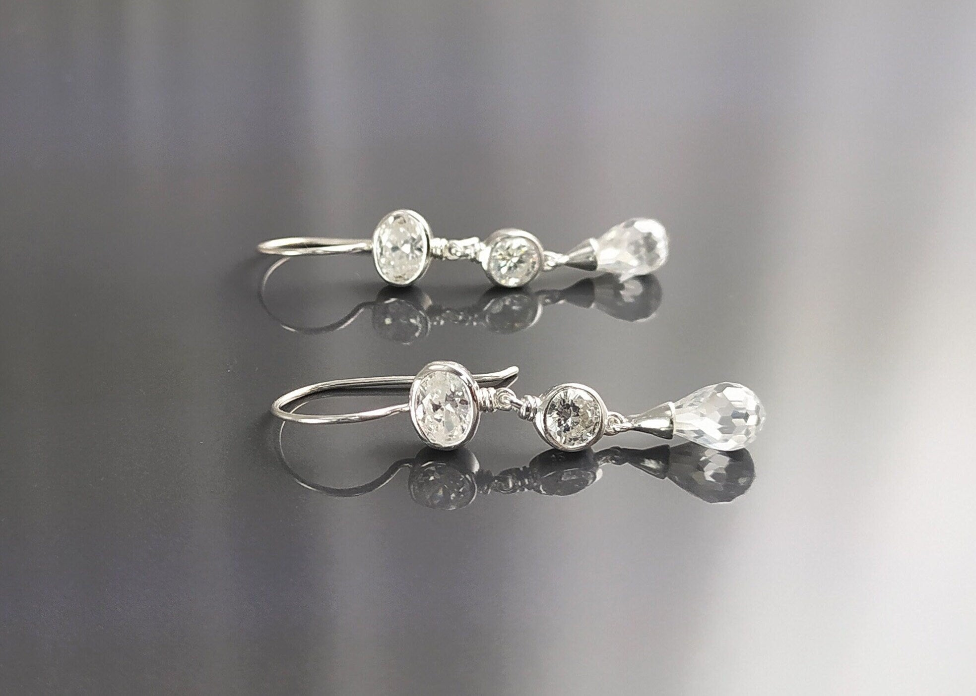 Une paire de boucles d'oreilles pendantes en argent avec trois pierres de cristal transparentes. La pierre du haut est de forme ovale, la pierre du milieu est ronde et la pierre du bas est en forme de goutte.