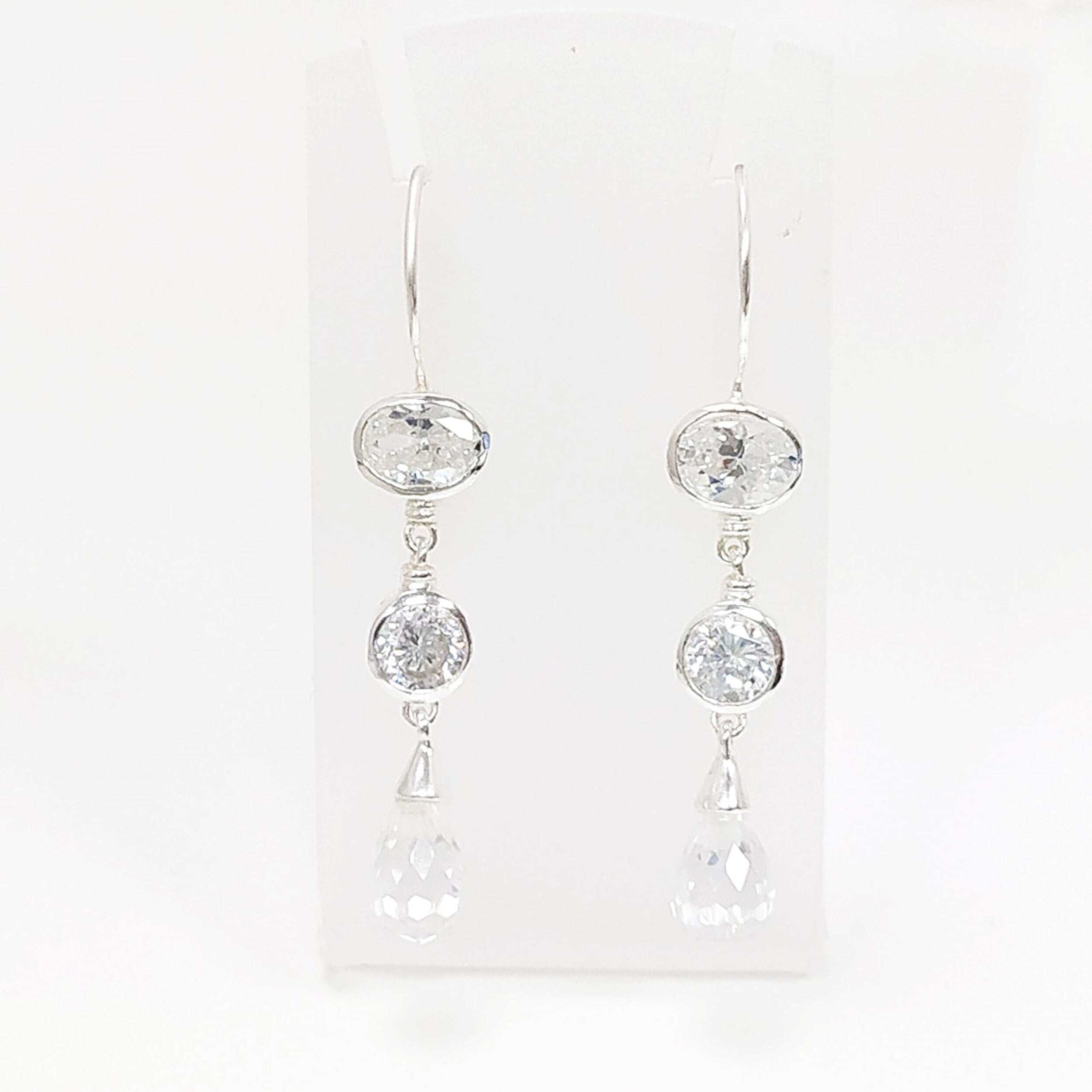 Une paire de boucles d'oreilles pendantes en argent avec trois pierres de cristal claires sur chaque boucle d'oreille. La pierre du haut est de forme ovale, la pierre du milieu est ronde et la pierre du bas est en forme de goutte.