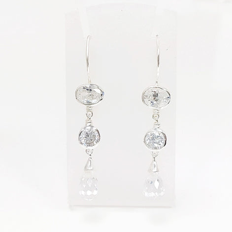 Une paire de boucles d'oreilles pendantes en argent avec trois pierres de cristal claires sur chaque boucle d'oreille. La pierre du haut est de forme ovale, la pierre du milieu est ronde et la pierre du bas est en forme de goutte.