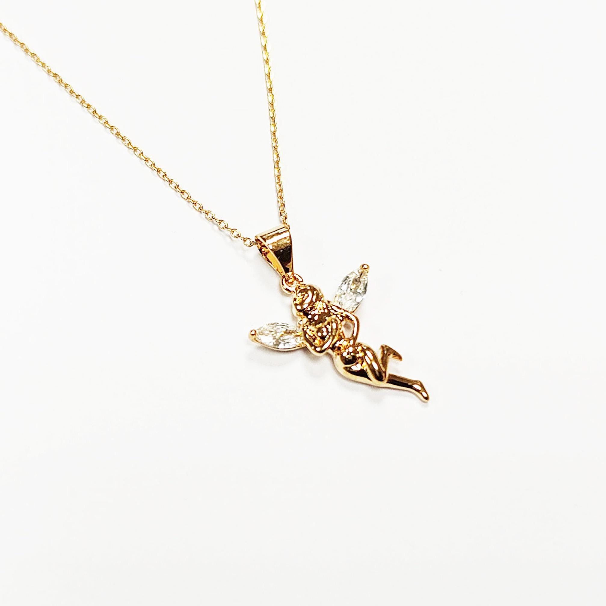 Un collier doré avec un pendentif en forme de fée. La fée a des ailes claires en forme de gemmes et est en position de vol. La chaîne du collier est délicate et dorée, sur fond blanc.