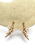 Boucles d'oreilles dorées en forme de fée avec des ailes en cristal transparent. Les boucles d'oreilles présentent un design détaillé d'une figurine de fée. Les boucles d'oreilles sont présentées sur une surface beige texturée, sur fond blanc.