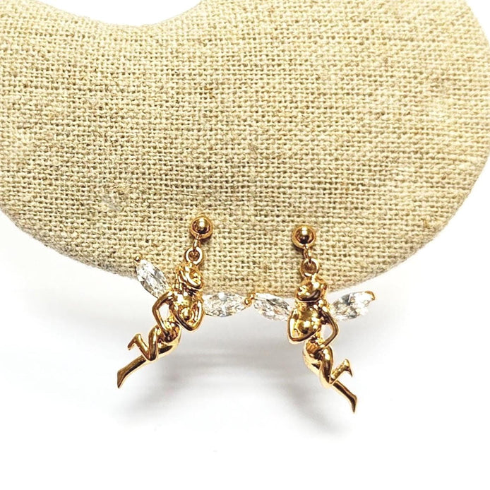 Boucles d'oreilles dorées en forme de fée avec des ailes en cristal transparent. Les boucles d'oreilles présentent un design détaillé d'une figurine de fée. Les boucles d'oreilles sont présentées sur une surface beige texturée, sur fond blanc.
