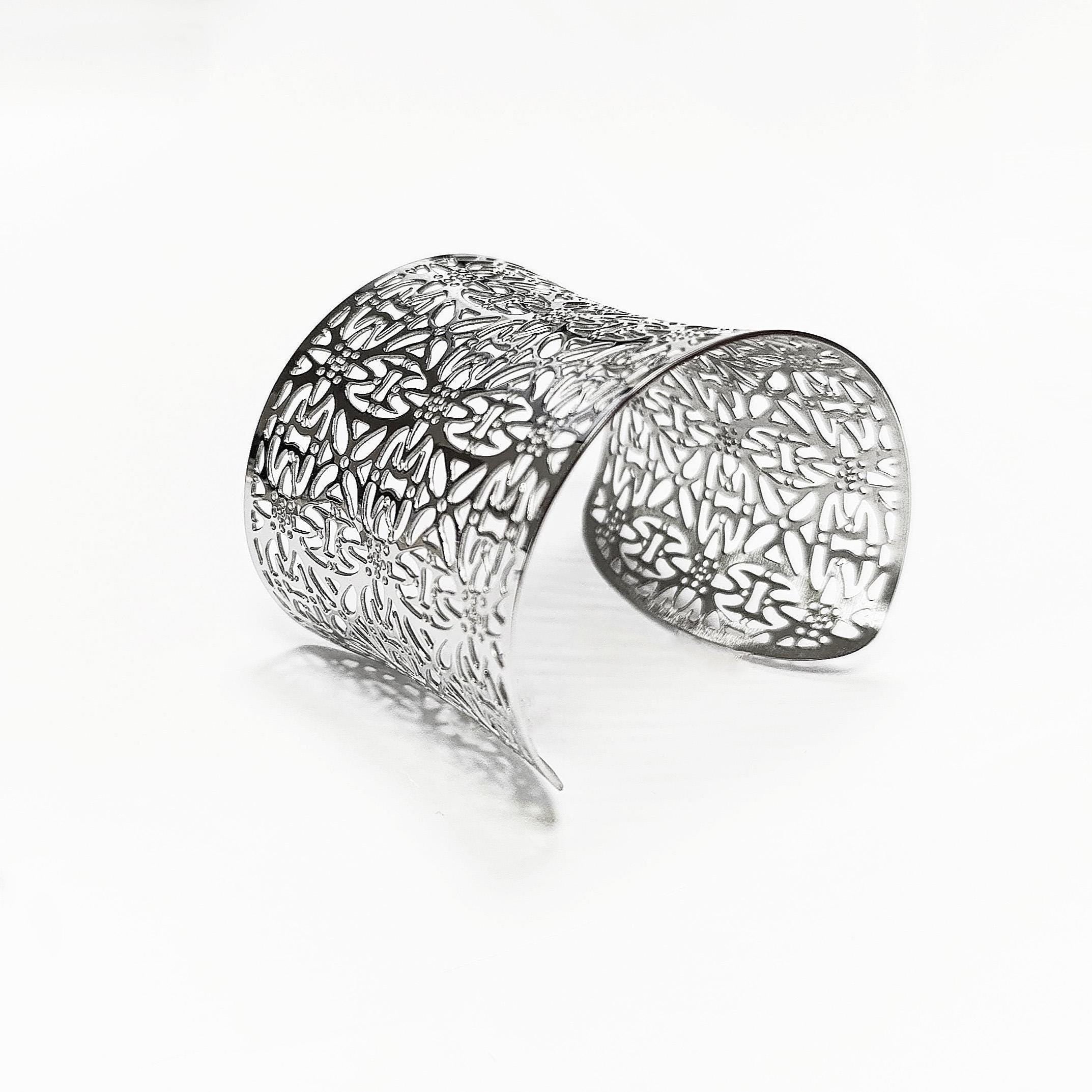 Angle view — A silver cuff bracelet with an openwork floral design. The wide bracelet curves gracefully, showcasing intricate cut-out patterns. The metal reflects light, creating highlights and shadows. The bracelet is a statement piece, suitable for various occasions.

Brand: KRAMIKE
Type of jewelry: cuff / statement armor cuff
Metal: stainless steel
Color: silver-tone
Style: Filigree floral lace / modern boho
Diameter: 66 mm
Width: 60 mm
Ships from: France