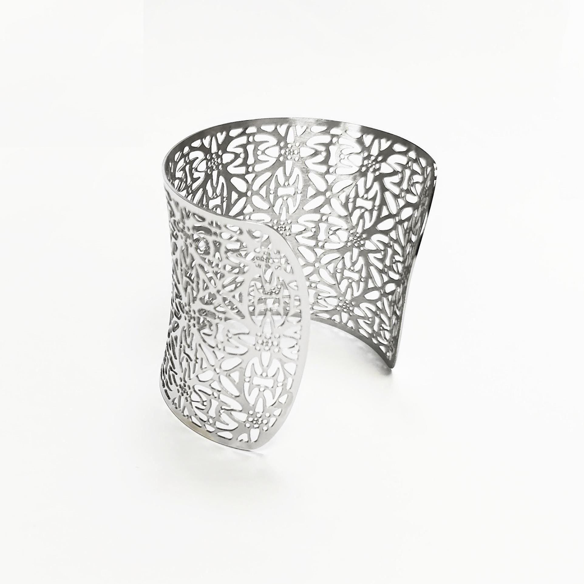 A silver-colored open cuff bracelet with a floral cutout design. The wide bracelet curves around, showcasing intricate patterns. The openwork design allows light to pass through, creating a delicate and airy aesthetic. This piece is a statement accessory.

Brand: KRAMIKE
Type of jewelry: cuff / statement armor cuff
Metal: stainless steel
Color: silver-tone
Style: Filigree floral lace / modern boho
Diameter: 66 mm
Width: 60 mm
Ships from: France