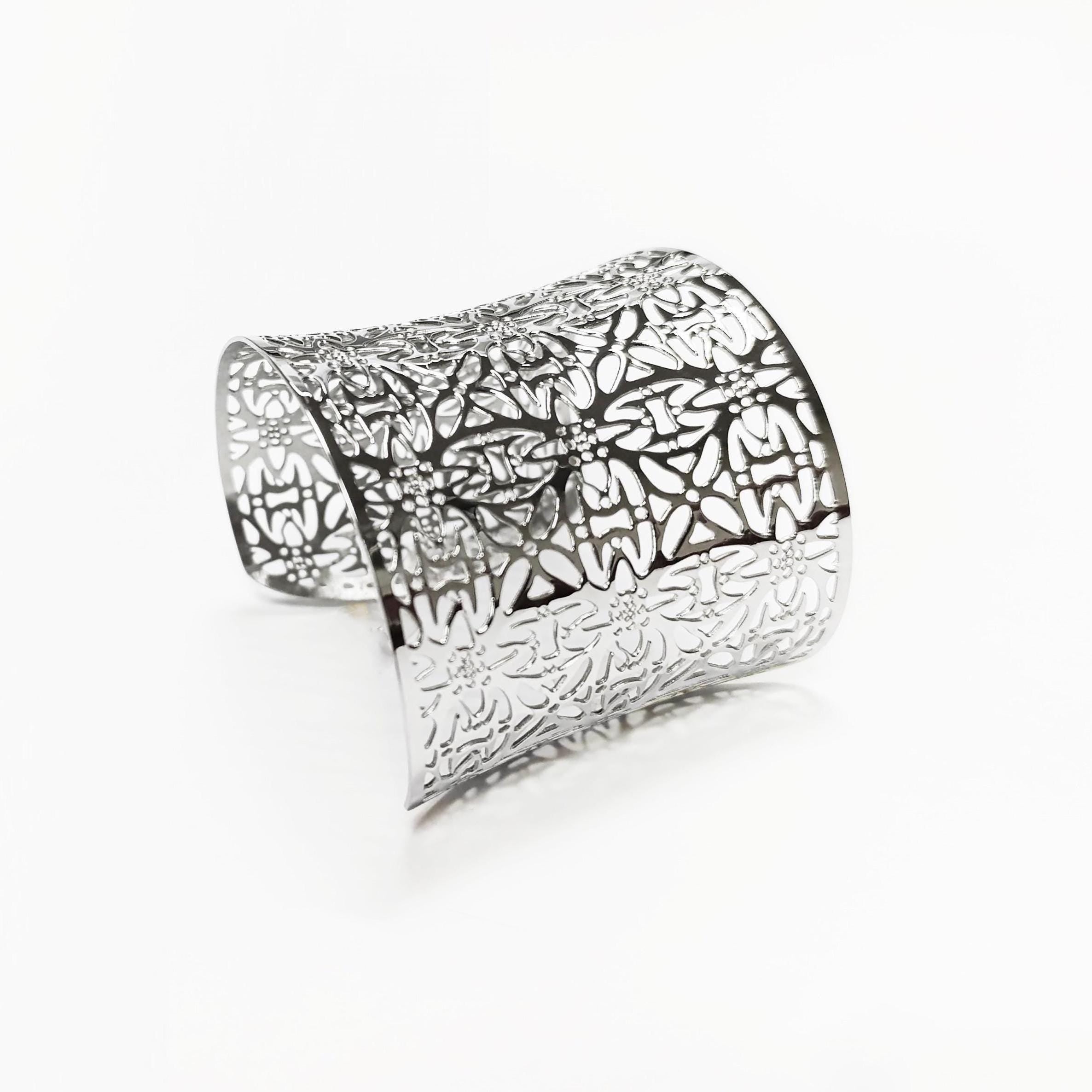 Angle view — Silver cuff bracelet with a floral pattern. The wide bracelet has a curved shape and is made of a shiny, reflective metal. The openwork design allows light to pass through, creating a delicate and intricate look.

Brand: KRAMIKE
Type of jewelry: cuff / statement armor cuff
Metal: stainless steel
Color: silver-tone
Style: Filigree floral lace / modern boho
Diameter: 66 mm
Width: 60 mm
Ships from: France