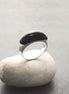Peut inclure: Une bague en argent avec une pierre ovale noire. La bague est posée sur un rocher blanc.