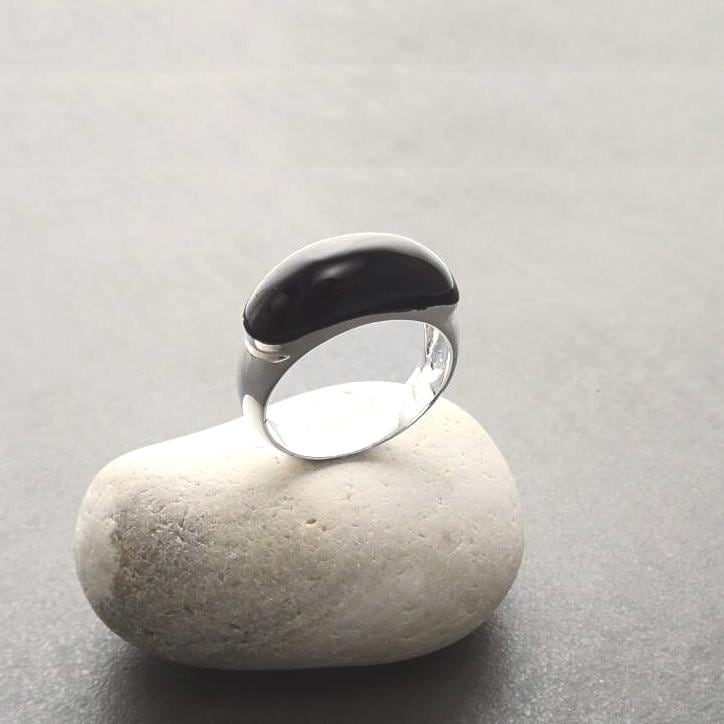Peut inclure: Une bague en argent avec une pierre ovale noire. La bague est posée sur un rocher blanc.