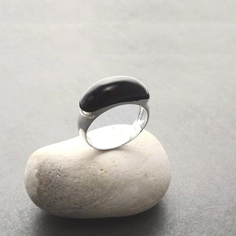 Peut inclure: Une bague en argent avec une pierre ovale noire. La bague est posée sur un rocher blanc.