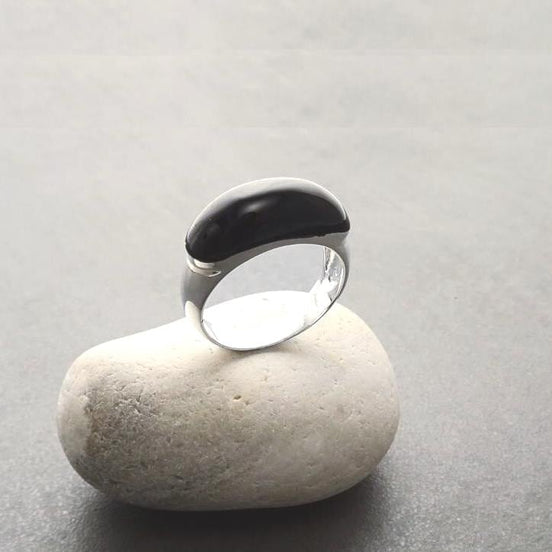 Peut inclure: Une bague en argent avec une pierre ovale noire. La bague est posée sur un rocher blanc.