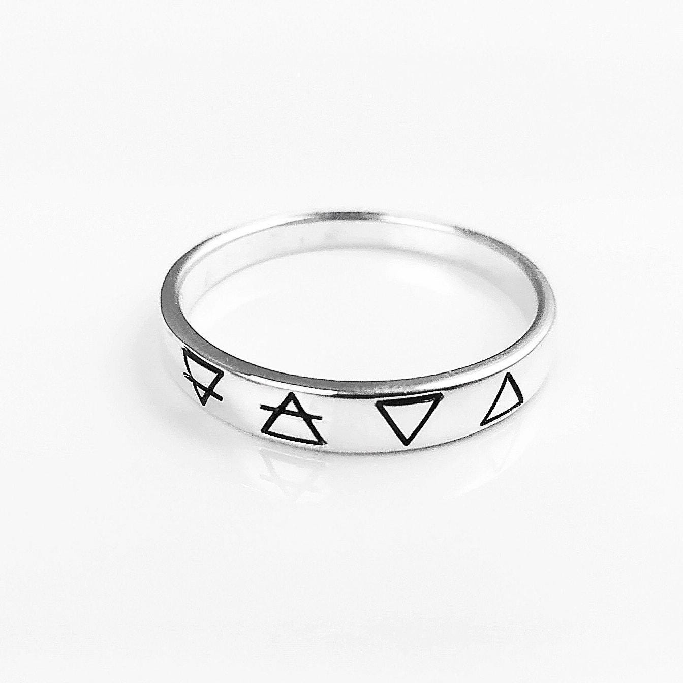 Une bague en argent avec quatre symboles gravés : un cercle avec une ligne qui le traverse, un triangle, un triangle inversé et un triangle avec une ligne qui le traverse.
VU D'en haut.
Bijou artisanal durable. Fait main local. Boutique de confiance recommandée.