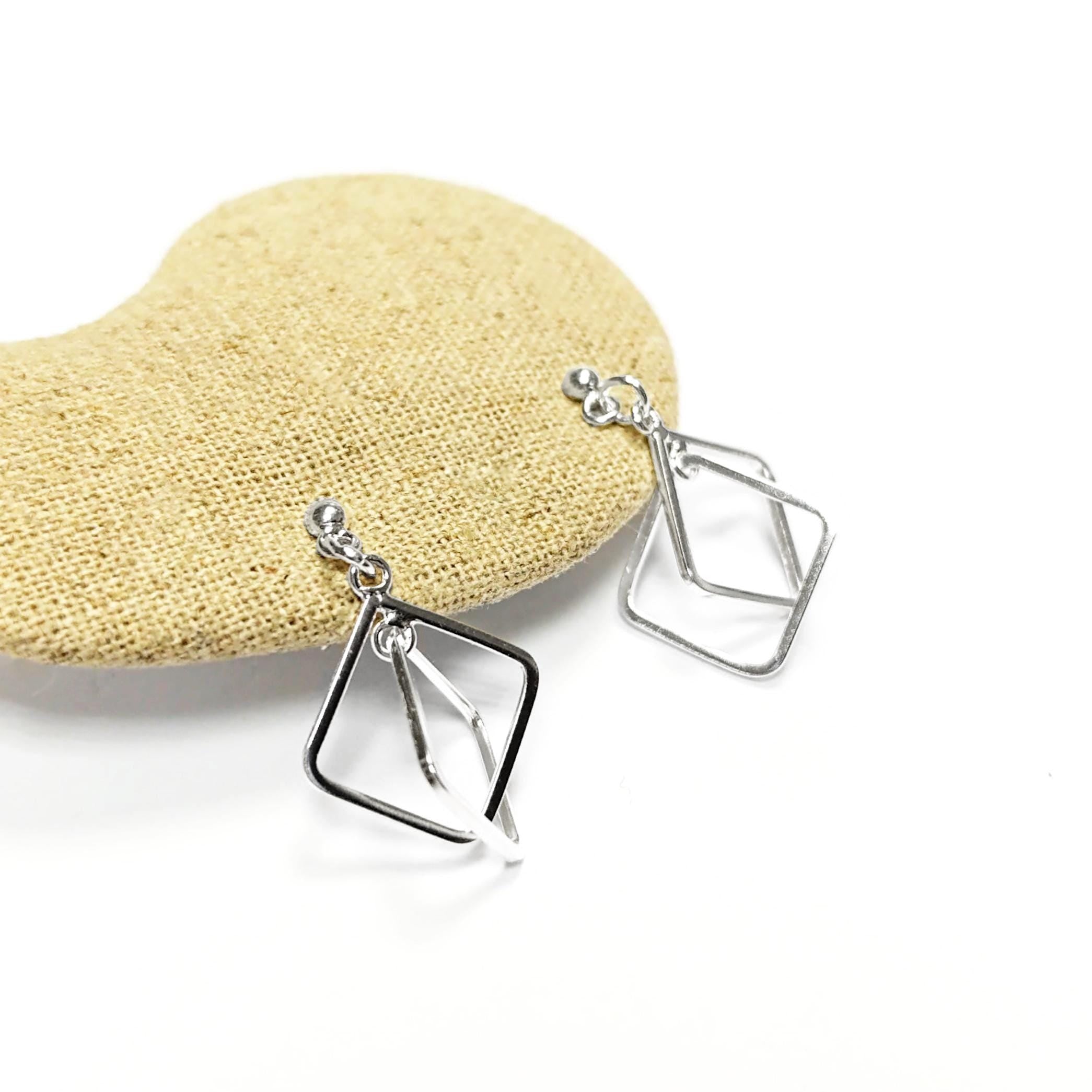 Peut inclure: Une paire de boucles d'oreilles argentées au design géométrique. Chaque boucle d'oreille présente un petit clou et une forme de losange ouverte à double couche. Les boucles d'oreilles sont présentées sur une surface texturée beige sur fond blanc.