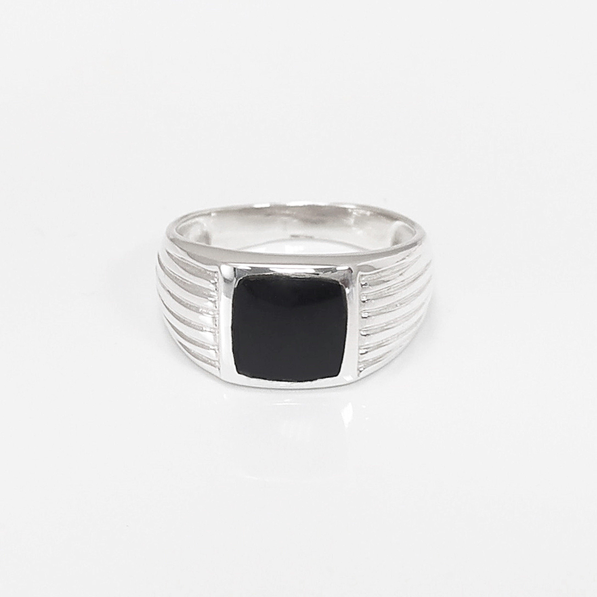 Peut inclure: Une bague en argent avec une pierre carrée noire sertie au centre. La bande de la bague a un design texturé.