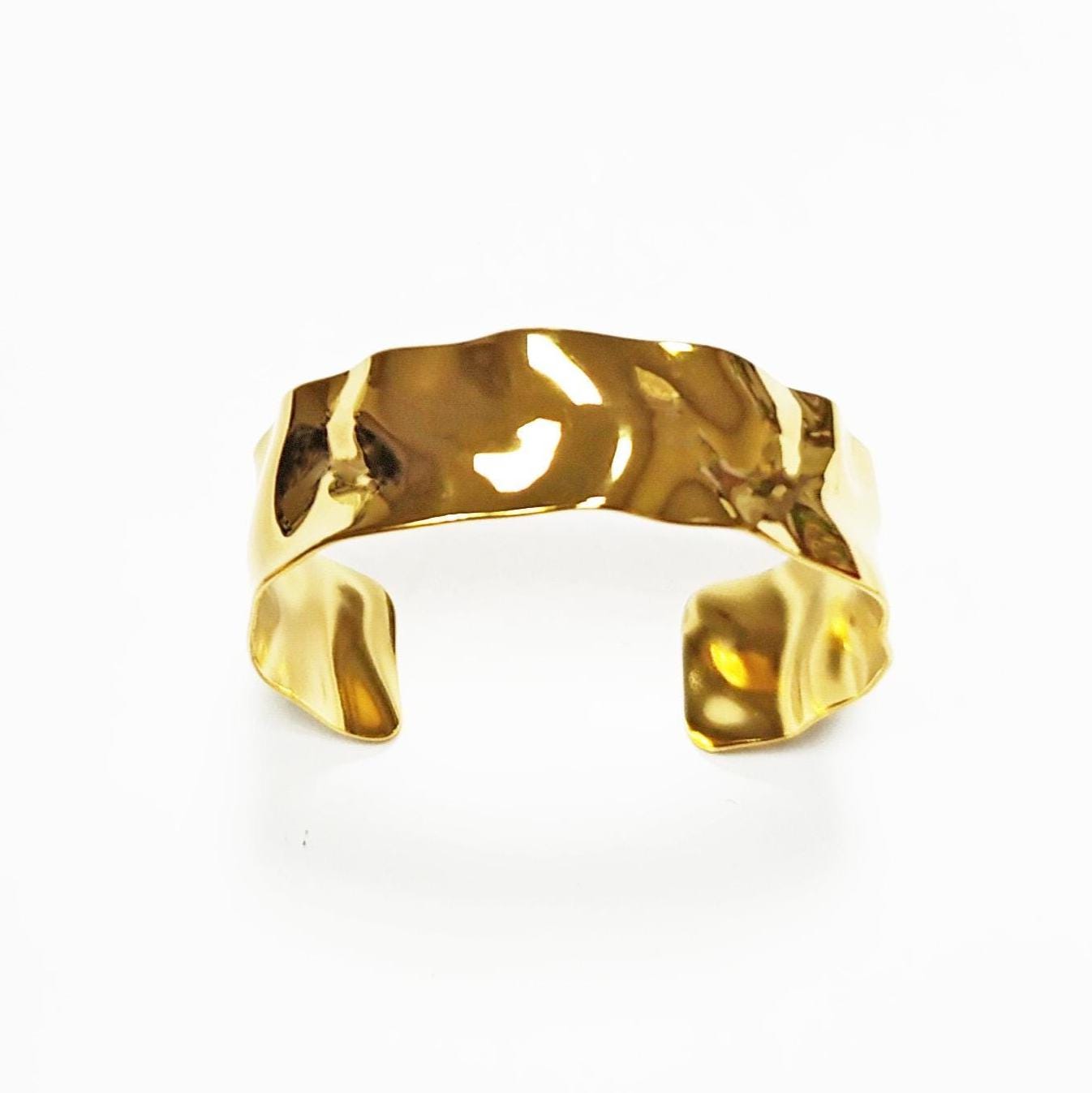 Peut inclure: Un bracelet manchette doré avec un aspect texturé et martelé. Le design ouvert présente une large bande de forme irrégulière et organique. Le bracelet est posé sur un fond blanc.