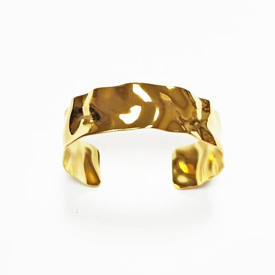 Peut inclure: Un bracelet manchette doré avec un aspect texturé et martelé. Le design ouvert présente une large bande de forme irrégulière et organique. Le bracelet est posé sur un fond blanc.