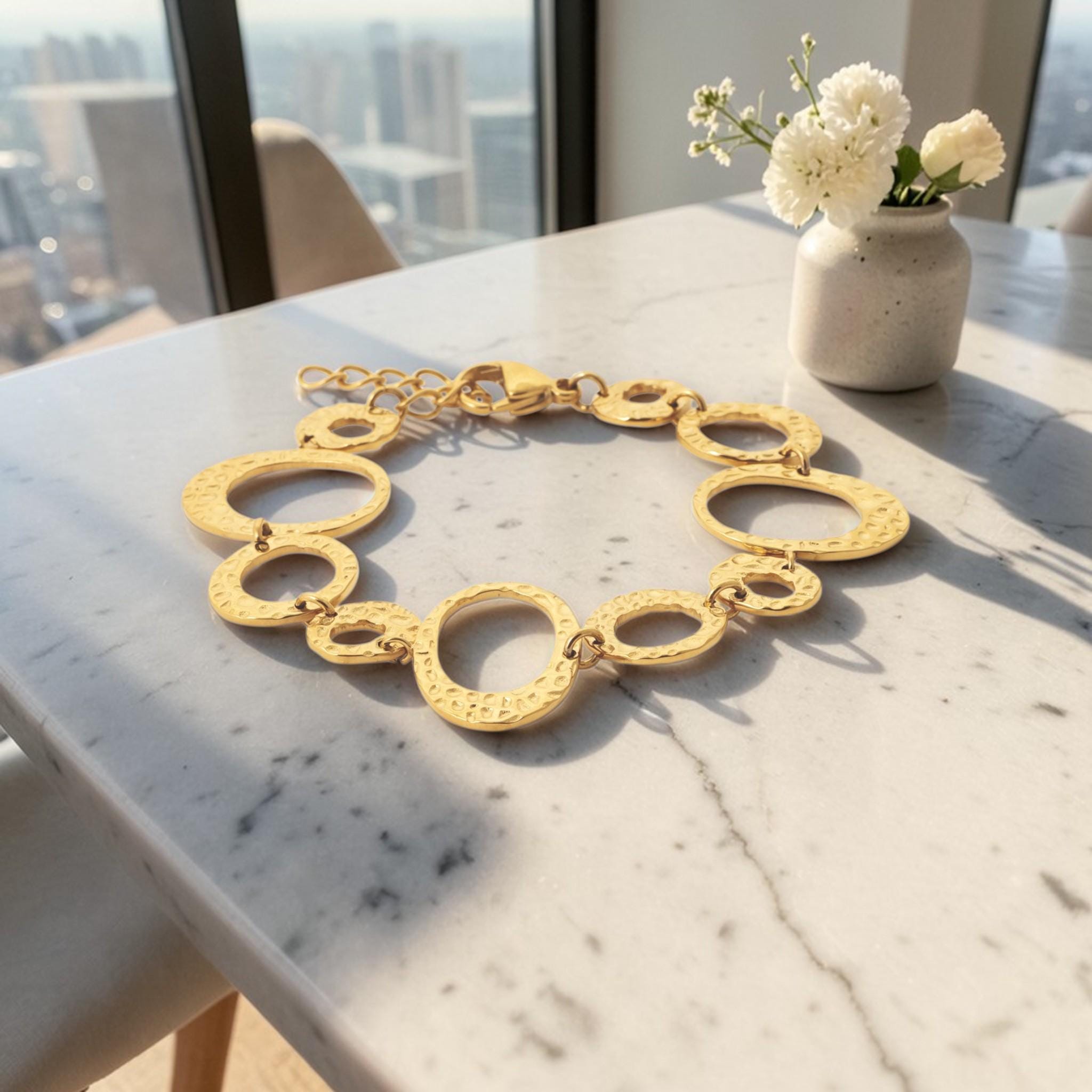 view of a geometric bracelet lay on a table
Product type: bracelet
Material:  18K Gold Filled, stainless steel base
Color: yellow gold
Style: statement, geometric, elegant, modern
Design details: hammered open circle and oval links, graduated sizes, textured finish, airy openwork design, lobster clasp with extender
Best for: statement styling, special occasions, everyday elevated wear, gifting, artistic jewelry lovers