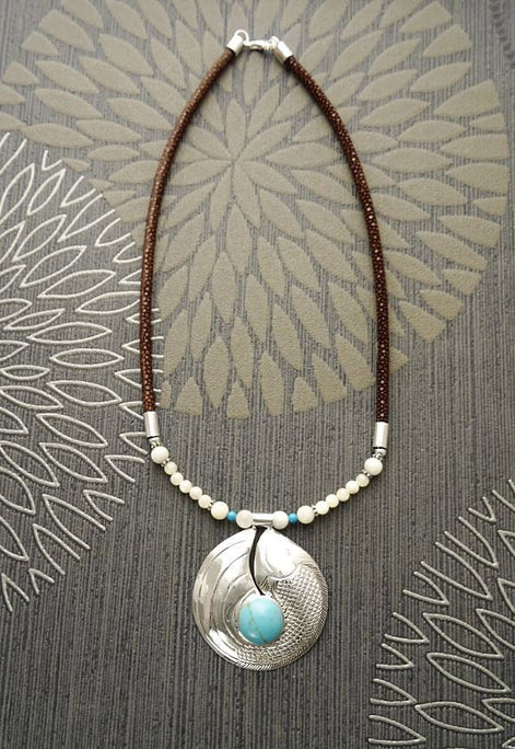 Peut inclure: Un collier en cordon de cuir marron avec un pendentif en argent orné d'une pierre turquoise et d'accents de perles blanches.
