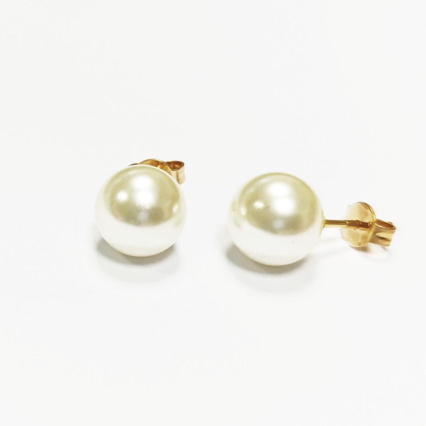 Une paire de boucles d'oreilles à clous en perles. Chaque boucle d'oreille présente une grosse perle ronde de couleur crème fixée sur une tige en or avec un fermoir papillon. Les boucles d'oreilles sont présentées sur une surface blanche.