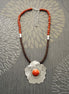 Peut inclure: Un collier avec un pendentif en forme de fleur argentée et une grande pierre de corail orange au centre. Le collier est composé de perles brunes et orange.