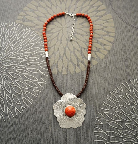 Peut inclure: Un collier avec un pendentif en forme de fleur argentée et une grande pierre de corail orange au centre. Le collier est composé de perles brunes et orange.