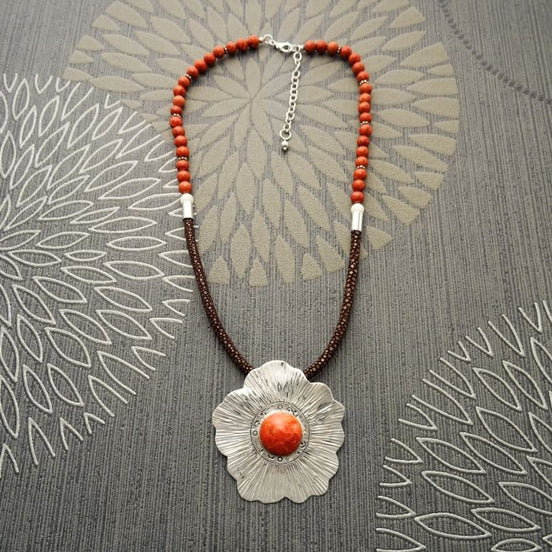 Peut inclure: Un collier avec un pendentif en forme de fleur argentée et une grande pierre de corail orange au centre. Le collier est composé de perles brunes et orange.