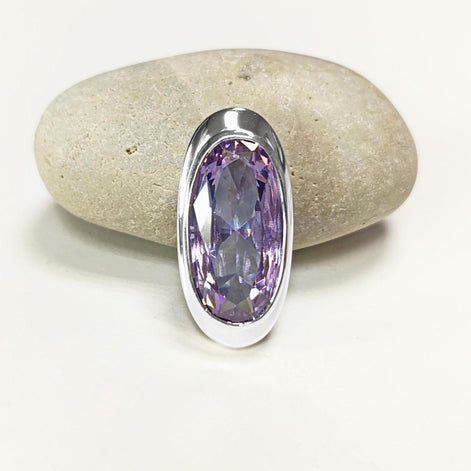 Peut inclure: Un pendentif argenté avec une grosse pierre précieuse ovale de couleur violet clair. La pierre est sertie dans une lunette argentée polie et repose sur une pierre grise claire. Le pendentif est présenté sur fond blanc.