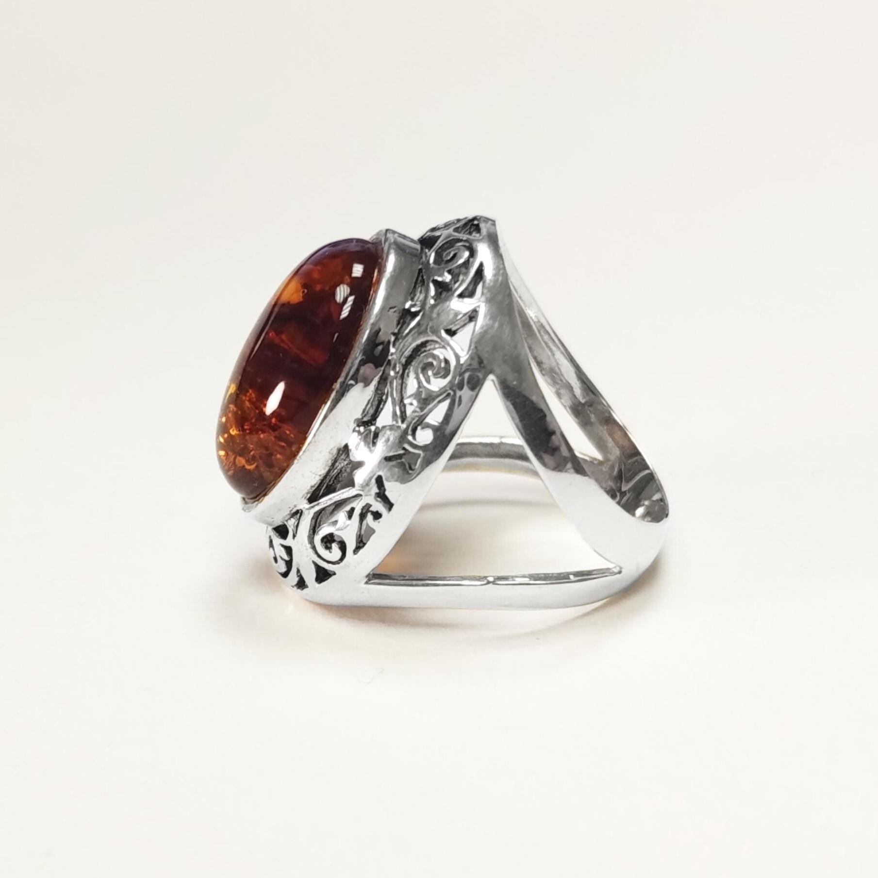 Peut inclure: Bague en argent ornée d'une pierre d'ambre ovale. La pierre d'ambre présente des couleurs chaudes et riches. L'anneau de la bague a un design ouvert avec des détails de filigrane décoratifs sur les côtés, ajoutant une touche élégante.
