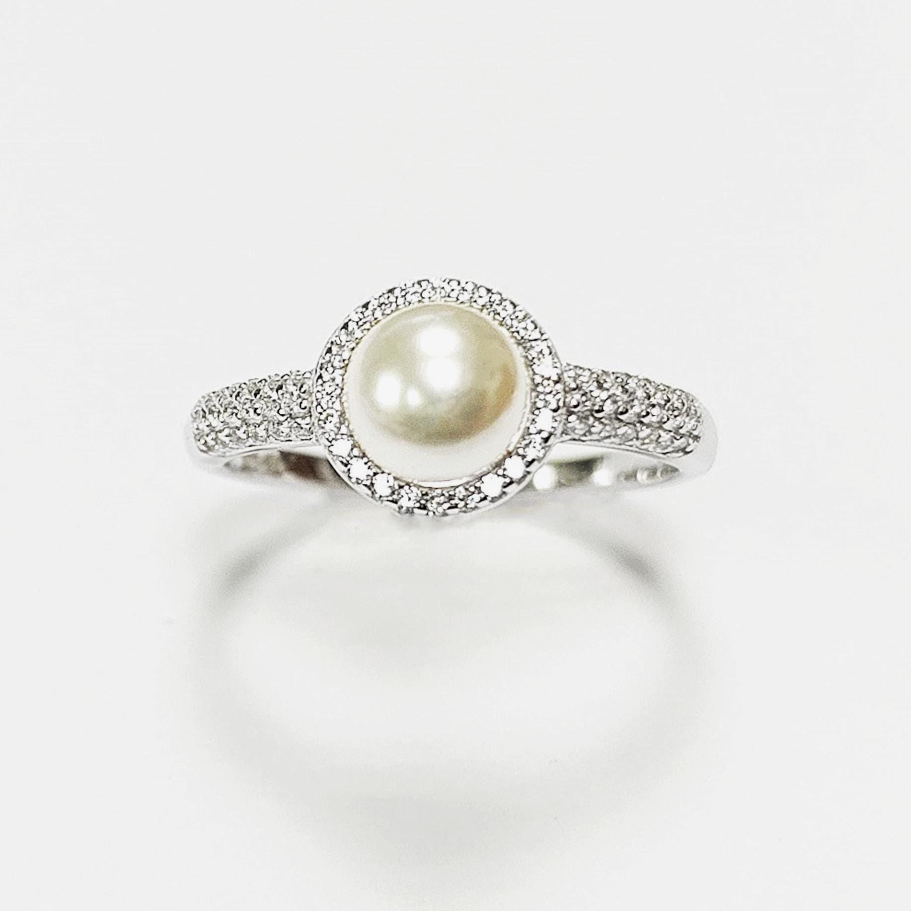 May include: A silver ring with a central, cream-coloured pearl surrounded by a halo of small, clear gemstones. The ring's band is also set with similar gemstones, creating a sparkling effect. The ring is displayed against a white background.