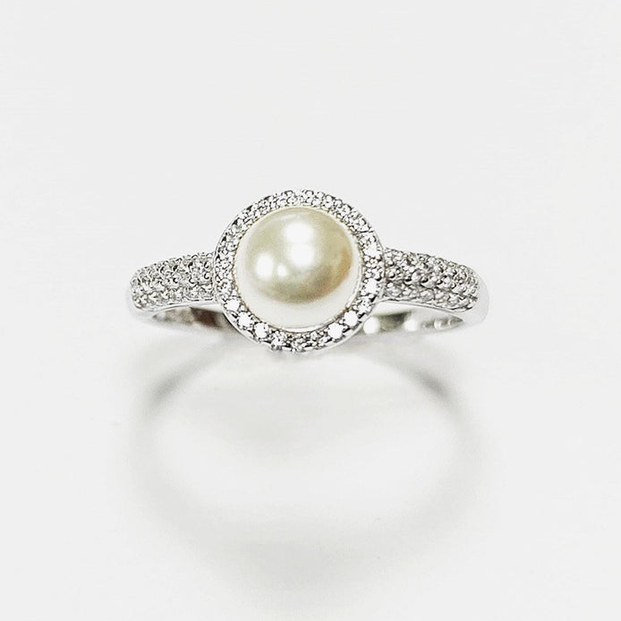 May include: A silver ring with a central, cream-coloured pearl surrounded by a halo of small, clear gemstones. The ring's band is also set with similar gemstones, creating a sparkling effect. The ring is displayed against a white background.