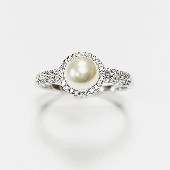 May include: A silver ring with a central, cream-coloured pearl surrounded by a halo of small, clear gemstones. The ring's band is also set with similar gemstones, creating a sparkling effect. The ring is displayed against a white background.