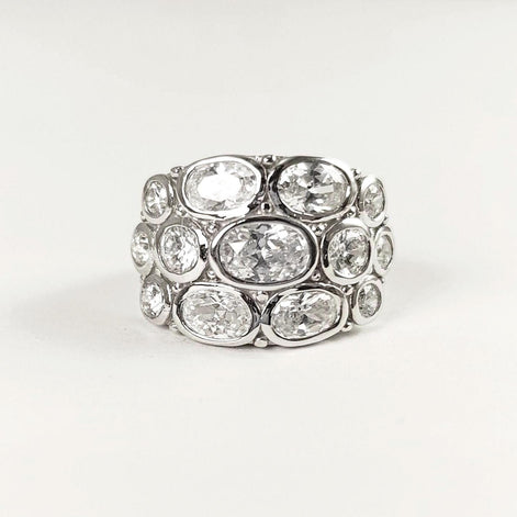 Peut inclure: Une large bague argentée ornée de plusieurs pierres précieuses ovales et transparentes. Les pierres sont disposées en couches, créant un design audacieux et scintillant. La bague est posée sur un fond blanc uni.
