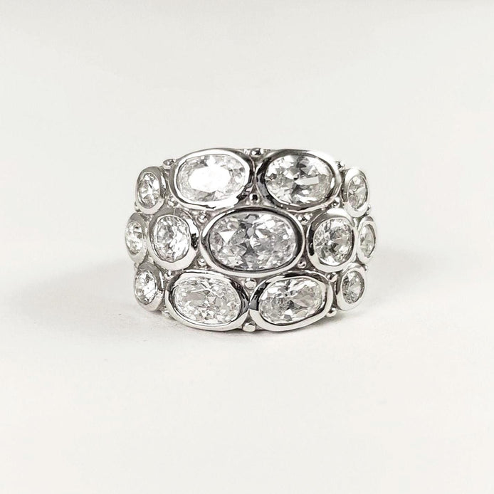 Peut inclure: Une large bague argentée ornée de plusieurs pierres précieuses ovales et transparentes. Les pierres sont disposées en couches, créant un design audacieux et scintillant. La bague est posée sur un fond blanc uni.