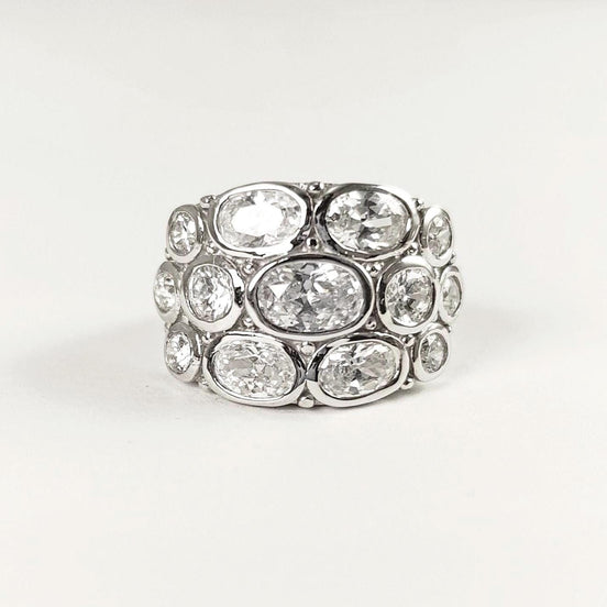 Peut inclure: Une large bague argentée ornée de plusieurs pierres précieuses ovales et transparentes. Les pierres sont disposées en couches, créant un design audacieux et scintillant. La bague est posée sur un fond blanc uni.