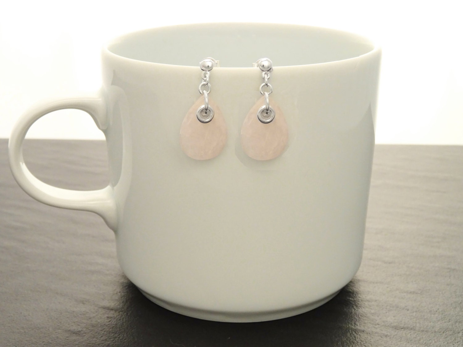 Une paire de boucles d'oreilles en forme de goutte d'eau en quartz rose clair et argent 925. Les boucles d'oreilles sont suspendues à une tasse en céramique blanche.