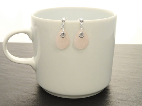 Une paire de boucles d'oreilles en forme de goutte d'eau en quartz rose clair et argent 925. Les boucles d'oreilles sont suspendues à une tasse en céramique blanche.