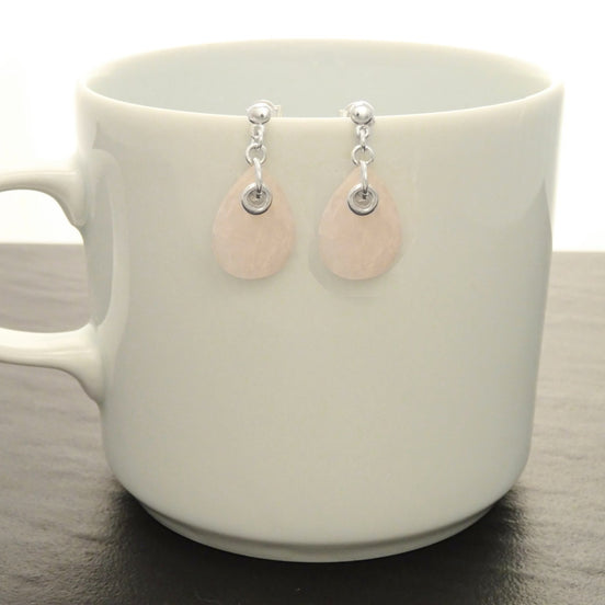 Une paire de boucles d'oreilles en forme de goutte d'eau en quartz rose clair et argent 925. Les boucles d'oreilles sont suspendues à une tasse en céramique blanche.